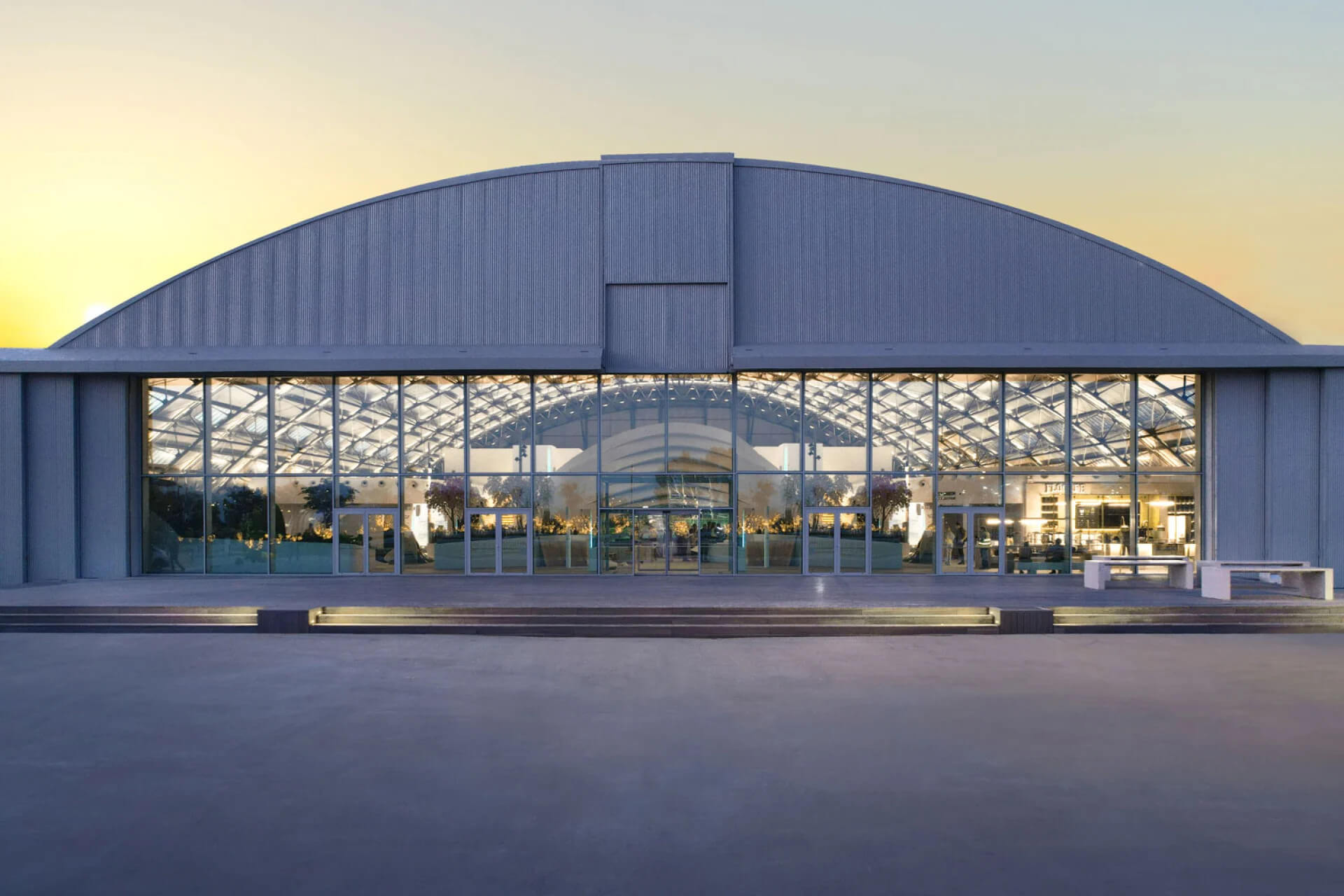 Θολωτό υπόστεγο Ellinikon Experience Center Κτίριο με υπόστεγο και γυάλινο τοίχο στην είσοδο. Πίσω φωτισμένος εσωτερικός χώρος.
