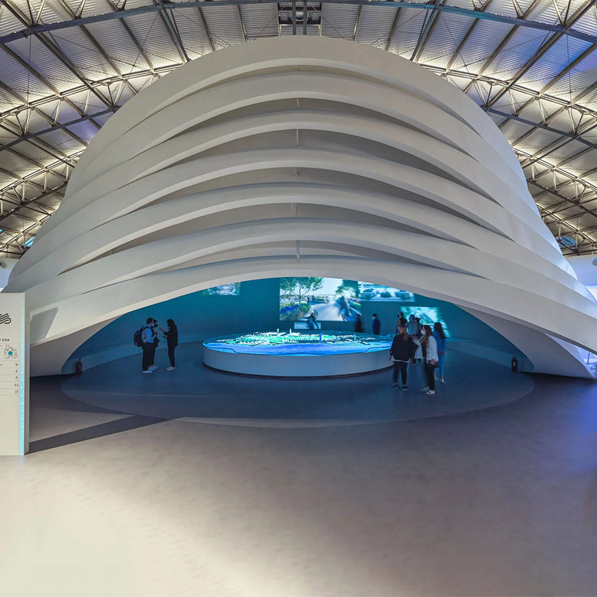 Εσωτερικό του Experience Center Λευκός μοντέρνος θόλος. Μέσα φωτισμένη μπλε μακέτα του πάρκου, γύρω επισκέπτες και στο βάθος οθόνες.