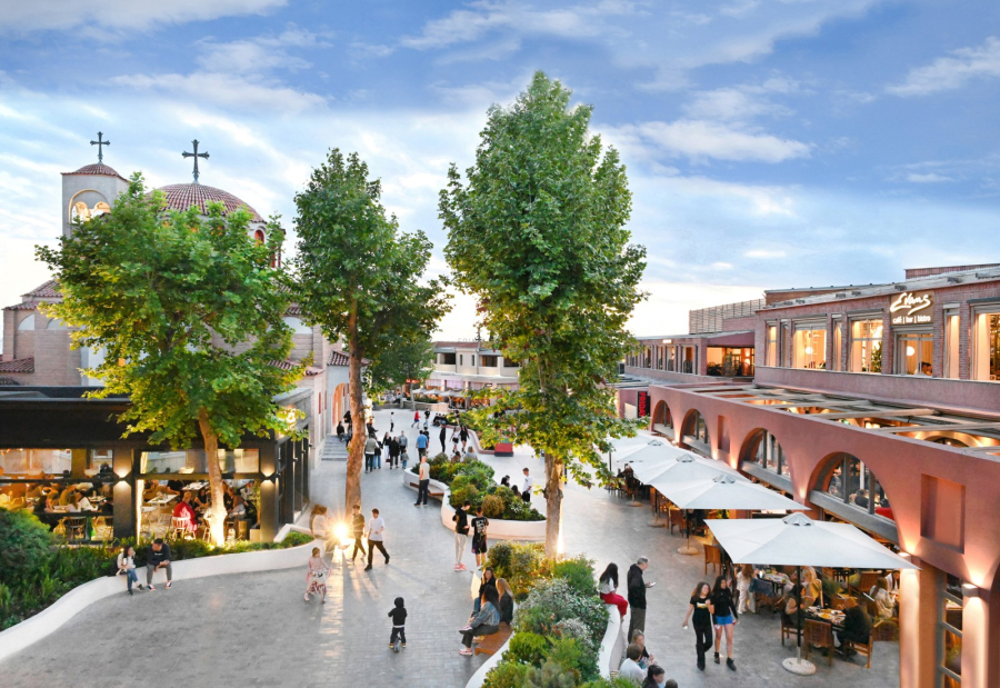 People walking in an outdoor corridor with trees and plants, restaurants to the left and right and a church at the back.