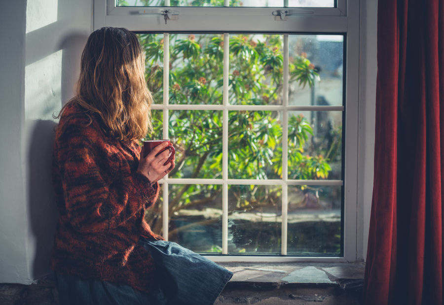 Χαμογελαστή νεαρή γυναίκα κάθεται σε καναπέ και κρατάει κούπα. Πίσω βιβλιοθήκη με φυτά και διακοσμητικά.