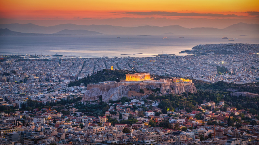Aerial view of Athens at dusk, the illuminated Parthenon at the center, buildings around it, the sea beyond and mountains in the distance.