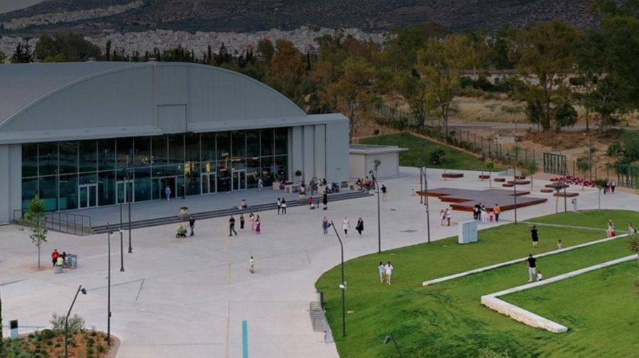 Γκρίζο υπόστεγο με γυάλινη πρόσοψη με εισόδους. Τριγύρω ανοικτός χώρος με γρασίδι. 