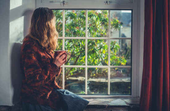 Χαμογελαστή νεαρή γυναίκα κάθεται σε καναπέ και κρατάει κούπα. Πίσω βιβλιοθήκη με φυτά και διακοσμητικά.